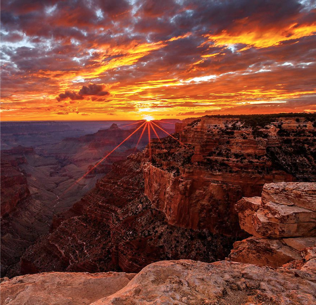 Dramatic canyon landscape at sunset with sun rays bursting over rock formations, vivid orange, red, and purple sky